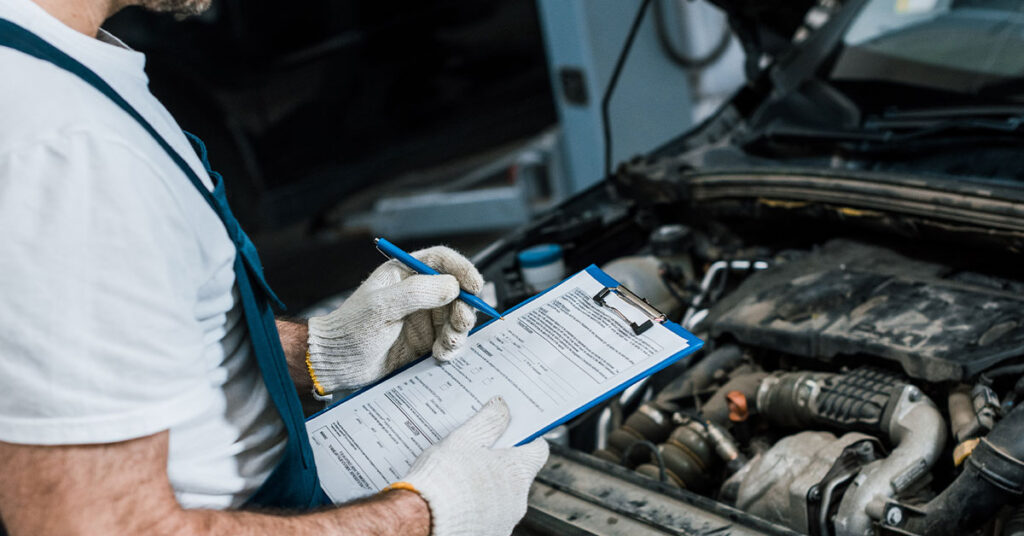 Mechanic holding a clipboard performing a safety check on a car with the bonnet raised exposing the engine bay.