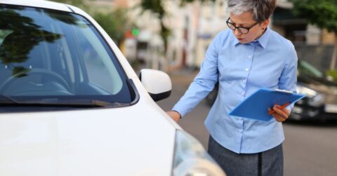 A woman holding a clipboard inspects a white car closely, likely evaluating its condition for appraisal or assessment purposes.