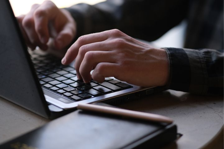 A close-up of a person typing on a black laptop keyboard, with a notebook and pen resting nearby on the table.