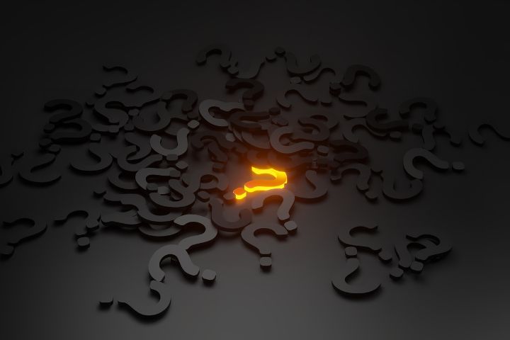 A glowing orange question mark stands out among a scattered pile of dark gray question marks on a dimly lit surface.