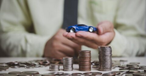 A person in a light green shirt holds a small blue toy car above a table covered with stacked and scattered coins, symbolizing car pricing or car financing decisions.